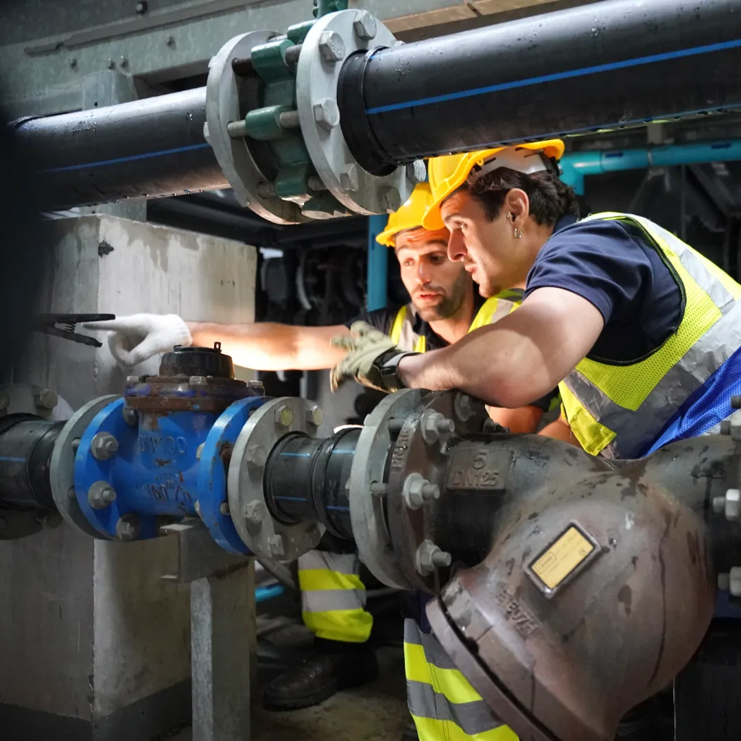 Two engineers in hi-vis inspecting valves and pipework in a plant room.