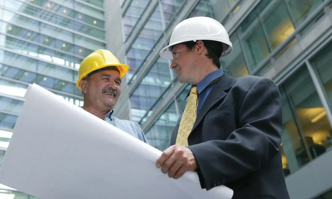 Two engineers in hard hats reviewing blueprints inside a modern glass office building.